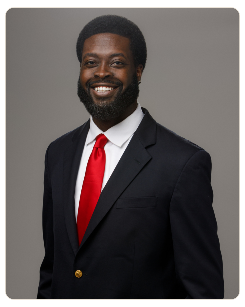 Ahmad Bryant Chief Executive Officer Smiling man in a navy suit with white shirt and red tie against a gray backdrop.