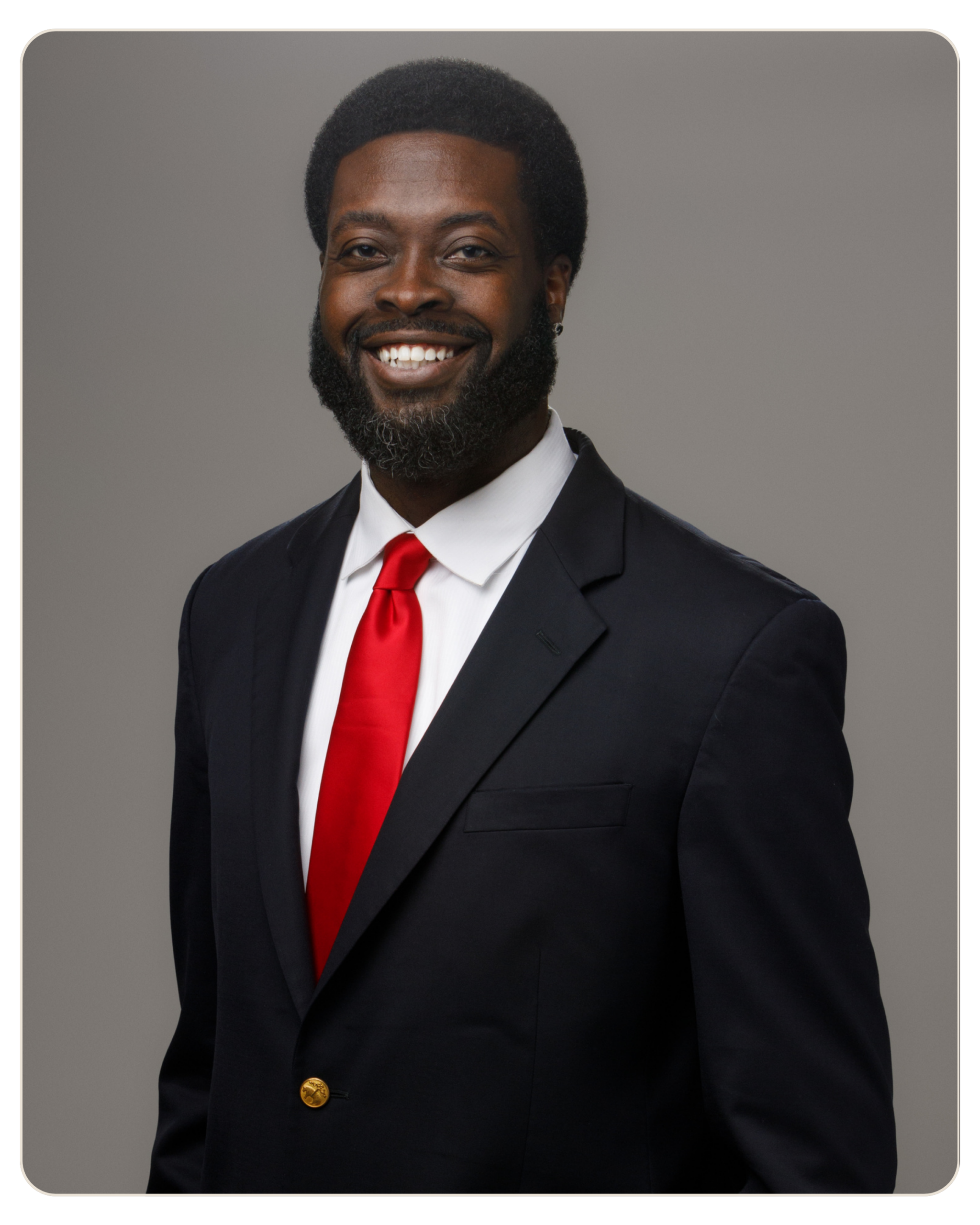 Ahmad Bryant Chief Executive Officer Smiling man in a navy suit with white shirt and red tie against a gray backdrop.