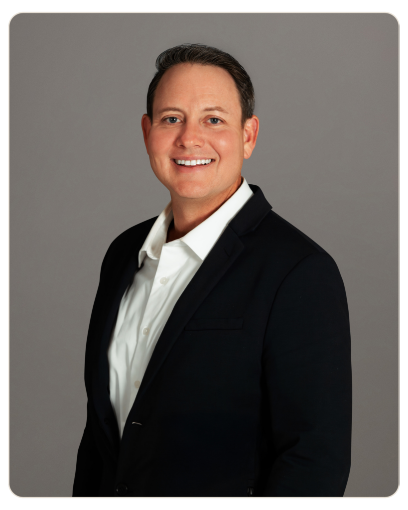 Flyland - Andrew Hooper Smiling man in a black suit and white shirt, head-and-shoulders portrait against a gray backdrop.