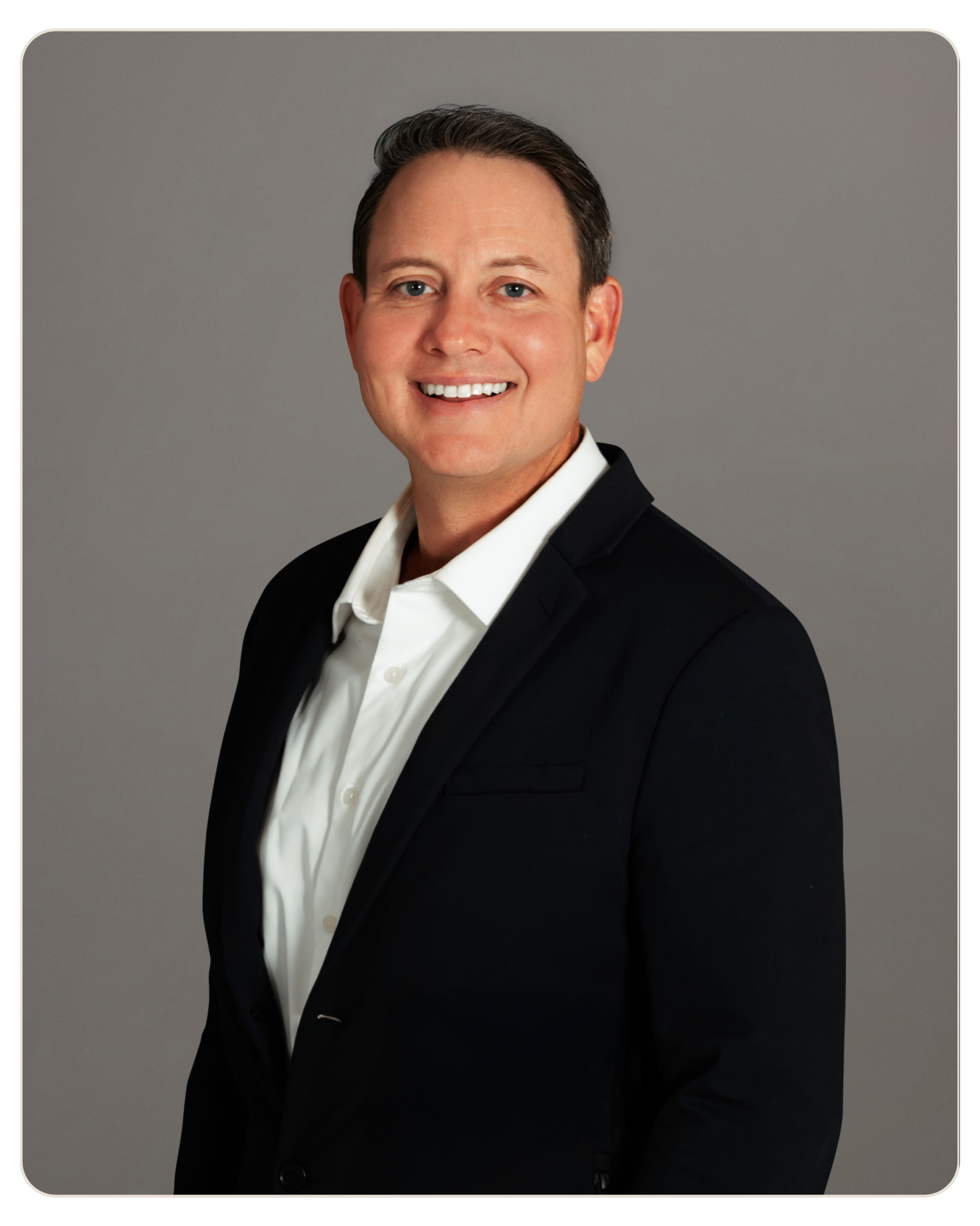 Flyland - Andrew Hooper Smiling man in a black suit and white shirt, head-and-shoulders portrait against a gray backdrop.